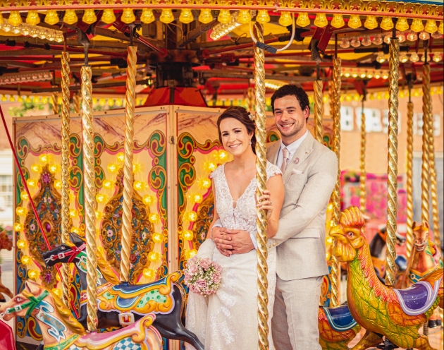 groom in cream suit, bride in slimline lace gown holding bouquet and sat on a carousel horse