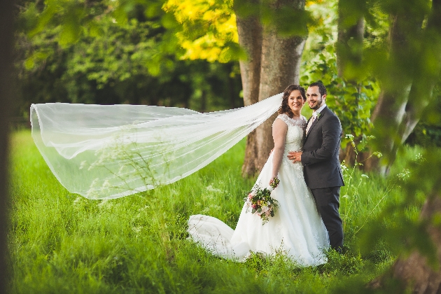 bride and groom in wedding attire standing on the grass surrounded by trees and bride's veil in the air