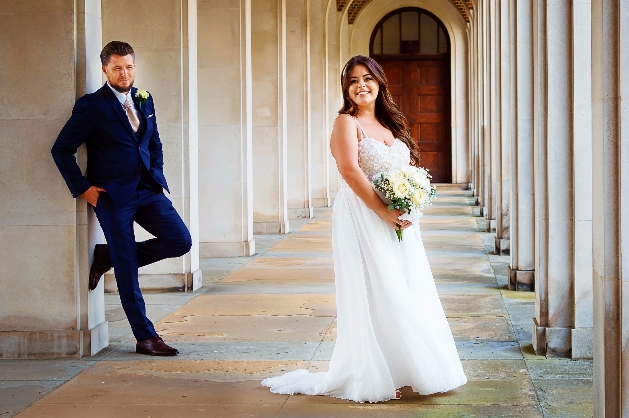 groom leaning on a wall looking at the bride standing near concrete pillars