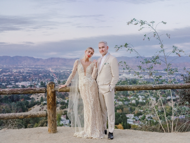 groom in cream suit next to bride in lace dress with plunging neckline and long train on a balcony