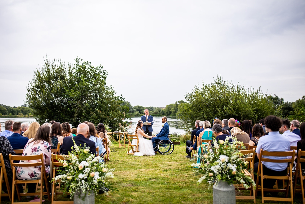 bride and groom saying their vows in front of a lake surrounded by rows of guests on chairs
