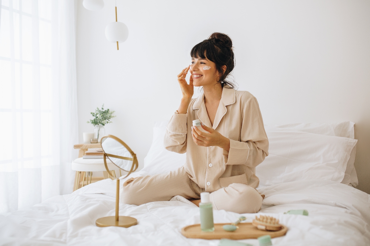 woman in her pjs on a bed applying moisturiser in a mirror