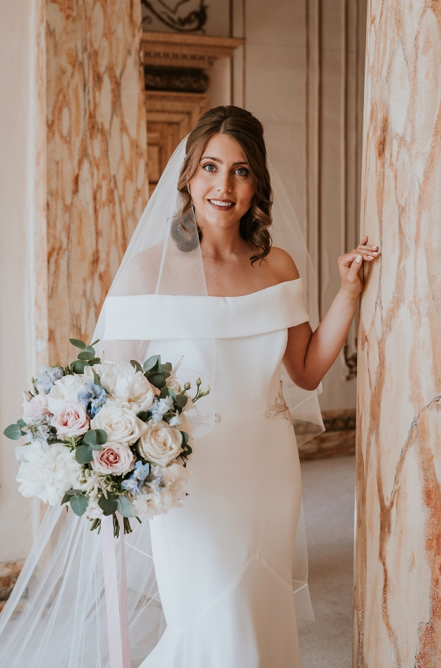 bride in off the shoulder dress and veil holding bouquet next to a marble pillar