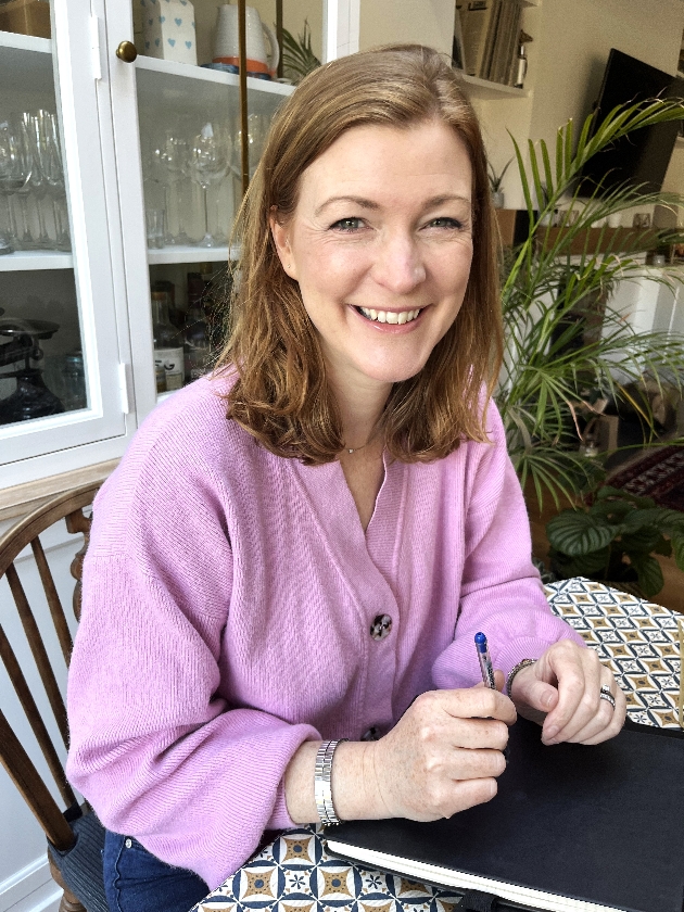 Celebrant suzie in a purple jumper in her kitchen sat at her table with a pad and pen