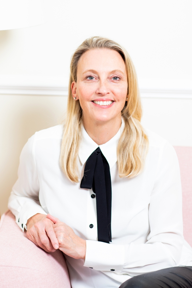 Blonde woman sat on pink chair wearing a white shirt with black bow detail